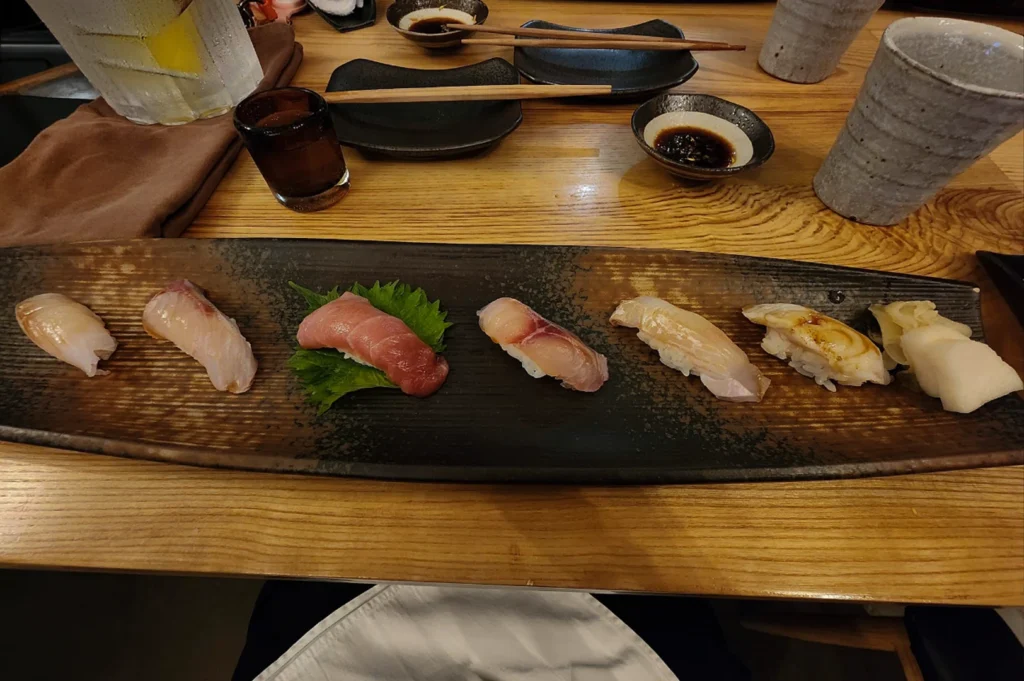 A high-angle, eye-level shot captures a long, dark, textured wooden serving platter centered on a light-colored wooden table, featuring an assortment of six pieces of nigiri sushi and a small pile of pickled ginger. From left to right, the nigiri consists of a white-fleshed fish, another piece of white fish, a bright red piece of tuna resting on a vibrant green shiso leaf, a third white-fleshed piece, a white piece brushed with a dark sauce, and finally, a small cluster of pickled ginger. Surrounding the platter on the table are various Japanese dining accessories, including two black ceramic dipping bowls filled with dark soy sauce, pairs of wooden chopsticks resting on black rectangular rests, a clear glass of water with ice, and a grey, textured ceramic cup. The overall scene reflects a refined, minimalist aesthetic typical of a high-end sushi dining experience.