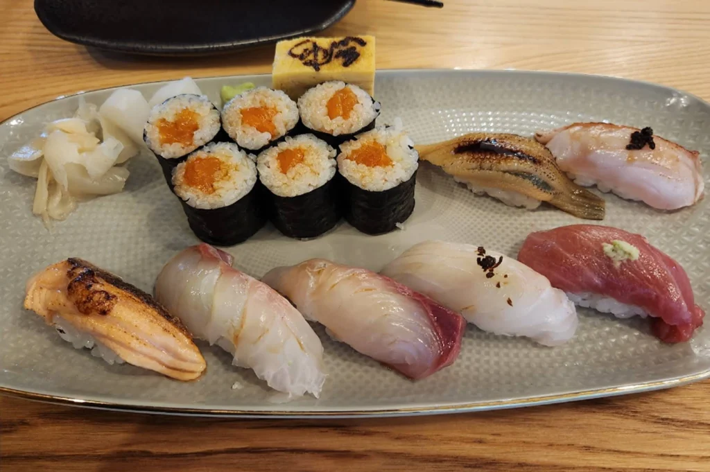 An oval plate filled with various sushi, including six pieces of maki rolls filled with orange roe, and seven pieces of nigiri featuring salmon, several types of white fish, tuna with wasabi, and unagi. A square of tamago (sweet omelet) with a branded mark sits at the top.