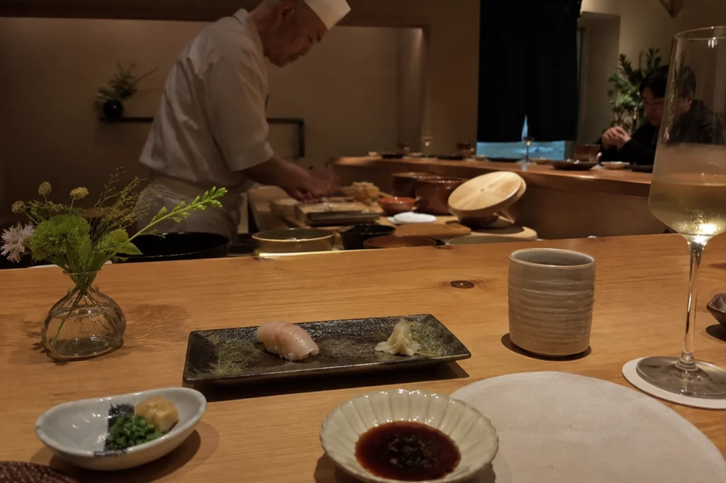 An eye-level close-up shot captures a piece of white fish nigiri with a touch of glaze, neatly placed on a dark, speckled rectangular ceramic plate alongside a small pile of ginger on a wooden sushi counter. In the foreground, a small side dish with green garnishes and a scalloped bowl of soy sauce rest next to a tall glass of white wine and a light-colored ceramic cup. In the background, an older sushi chef in a white uniform works diligently behind the counter, while a small clear glass vase holding delicate green flowers and a glimpse of other diners down the counter add to the warm, intimate atmosphere of the restaurant.