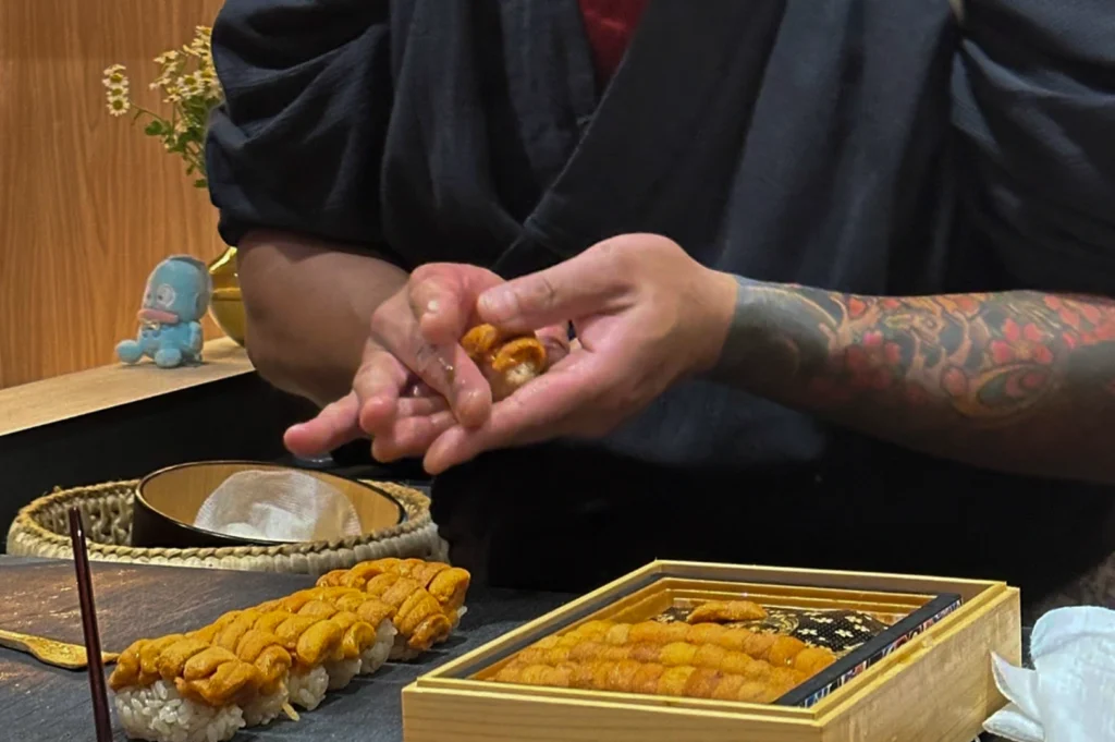 A close-up shot focuses on a sushi chef’s hands as they meticulously shape a piece of uni (sea urchin) nigiri. The chef wears a dark grey wrap-style uniform, and their left forearm features a vibrant, detailed tattoo with floral patterns. On a dark slate platter to the left, a row of pre-formed sushi rice topped with bright orange sea urchin sits ready, while a premium wooden box filled with rows of fresh uni rests on the counter to the right. A small blue cartoon figurine sits subtly in the soft-focused background.