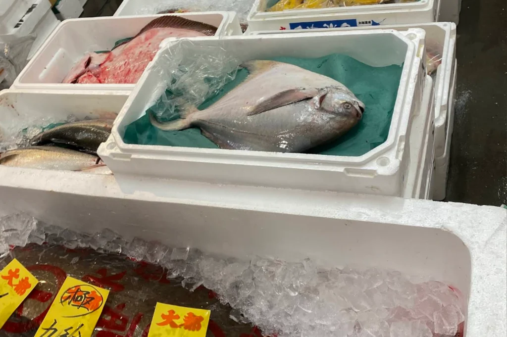 A high-angle view of several white styrofoam crates filled with fresh seafood at a fish market. The central crate prominently displays a large, silver pomfret fish resting on a piece of green paper, while other crates contain different varieties of fish packed in crushed ice. Bright yellow labels with Japanese characters are visible in the foreground, indicating the origin or type of the fresh catch.