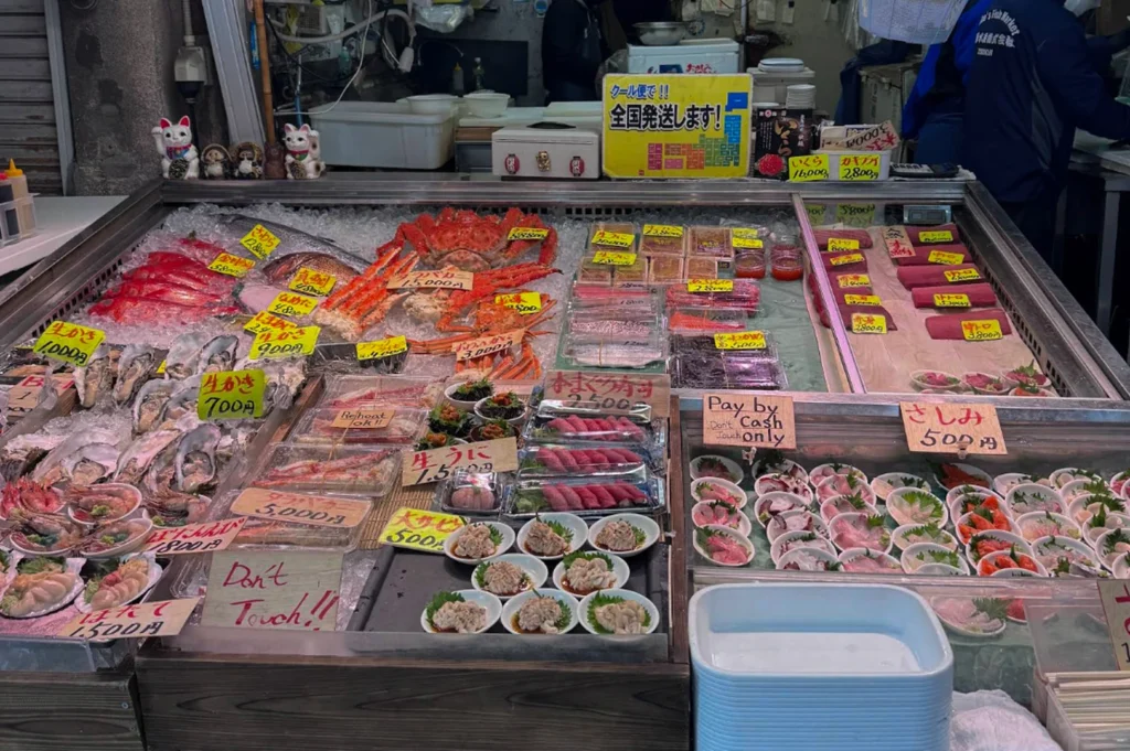 An overhead view of a large, rectangular market stall packed with various seafood on ice. On the left are bright red whole fish and fresh oysters. The center features a large whole king crab and several trays of sea urchin (Uni) and sliced tuna. The right side shows organized rows of pre-cut sashimi blocks. Small white plates of prepared snacks sit in the foreground. Several signs are visible, including one in English that reads, "Pay by Cash only" and "Don't Touch!!"