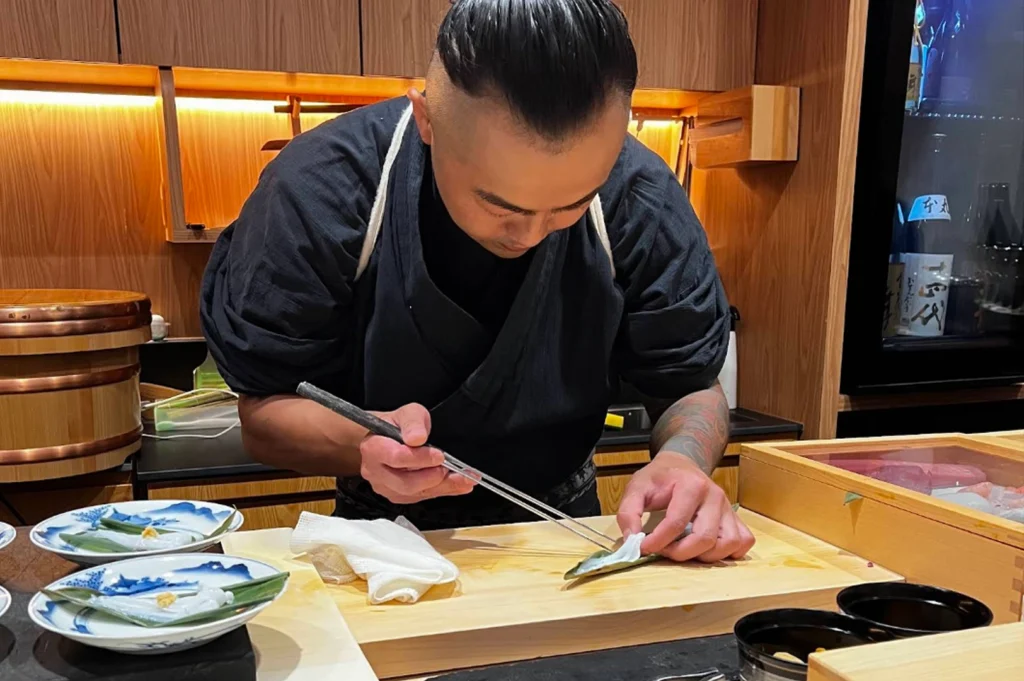 A focused shot of a sushi chef with a modern undercut hairstyle and a tattooed arm, dressed in a traditional dark samue or chef’s uniform. He is leaning over a light wood cutting board, using long, slender metal plating chopsticks to delicately arrange a piece of translucent white fish onto a small green leaf. In the foreground, several small, ornate blue and white ceramic plates are ready for service, while the background shows a well-organized wooden kitchen with professional tools.