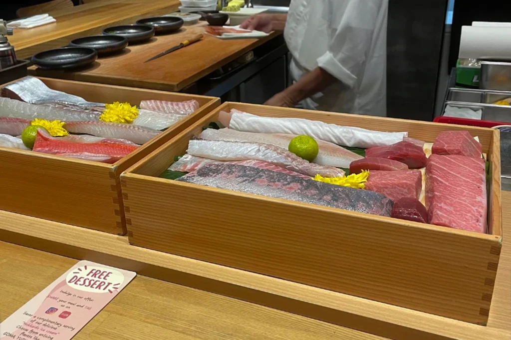 Two wooden boxes (neta bako) displayed on a sushi counter, showcasing high-quality cuts of raw fish ready for preparation. Visible items include marbled tuna (otoro), silver-skinned fish, and white fish, garnished with lime and yellow flowers. A chef is partially visible in the background.