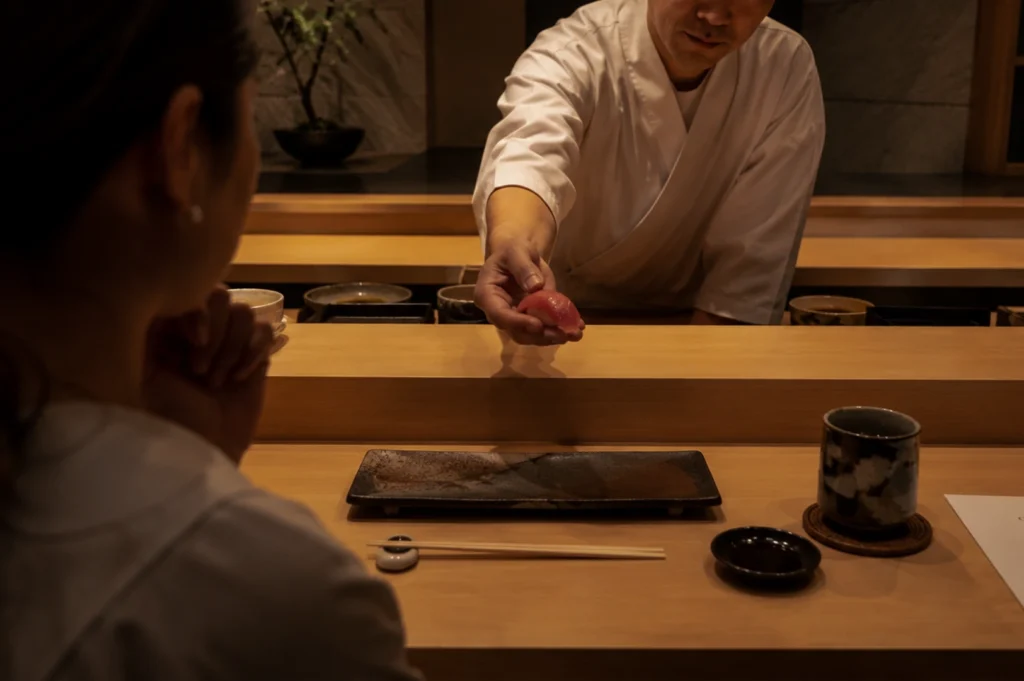 A close-up, eye-level shot shows a sushi chef leaning across a wide wooden counter to hand a piece of pink tuna nigiri to a guest. The chef, dressed in a traditional white chef's uniform, looks down thoughtfully as he extends his hand. In the foreground, the back of the guest's head and shoulder are visible as she watches intently. A dark, textured ceramic serving plate sits empty in front of her, flanked by a pair of wooden chopsticks on a ceramic rest, a small dish of soy sauce, and a rustic ceramic teacup.