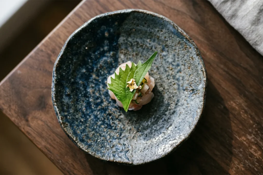 Gourmet Japanese appetizer featuring fresh sashimi topped with a green shiso leaf and edible gold leaf, served in a rustic ceramic bowl.