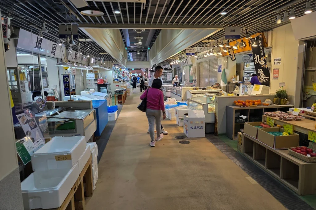 A perspective shot looking down a clean, modern indoor market hallway with a high ceiling featuring exposed slats and track lighting. The floor is a smooth, light-brown concrete. On the left, several white Styrofoam shipping boxes are stacked near a stall. On the right, a vendor sells fresh fruit, including bright oranges, red apples, and pineapples. A few shoppers are walking away from the camera toward the end of the bright corridor.
