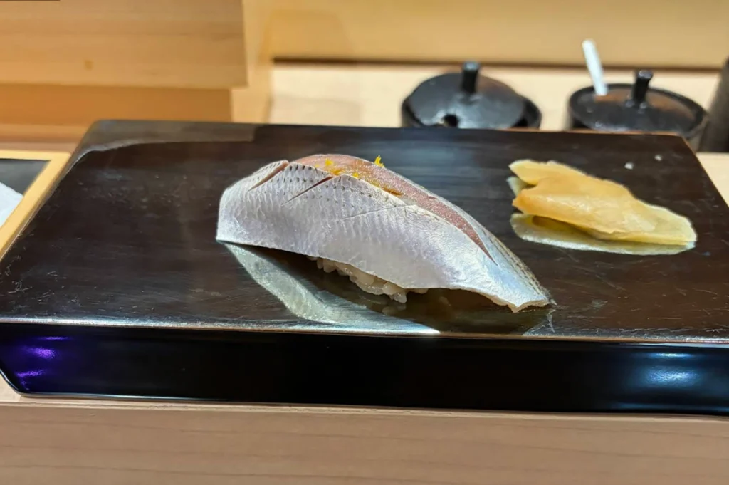 A close-up shot of a single piece of nigiri sushi presented on a sleek, reflective black rectangular platter. The sushi features a slice of silver-skinned fish, likely kohada, which has been precisely scored with diagonal cuts and garnished with a tiny pinch of yellow citrus zest. To the right of the sushi sits a small pile of pale, thinly sliced pickled ginger. The background is softly blurred, showing the light wood of the sushi counter and dark condiment containers.