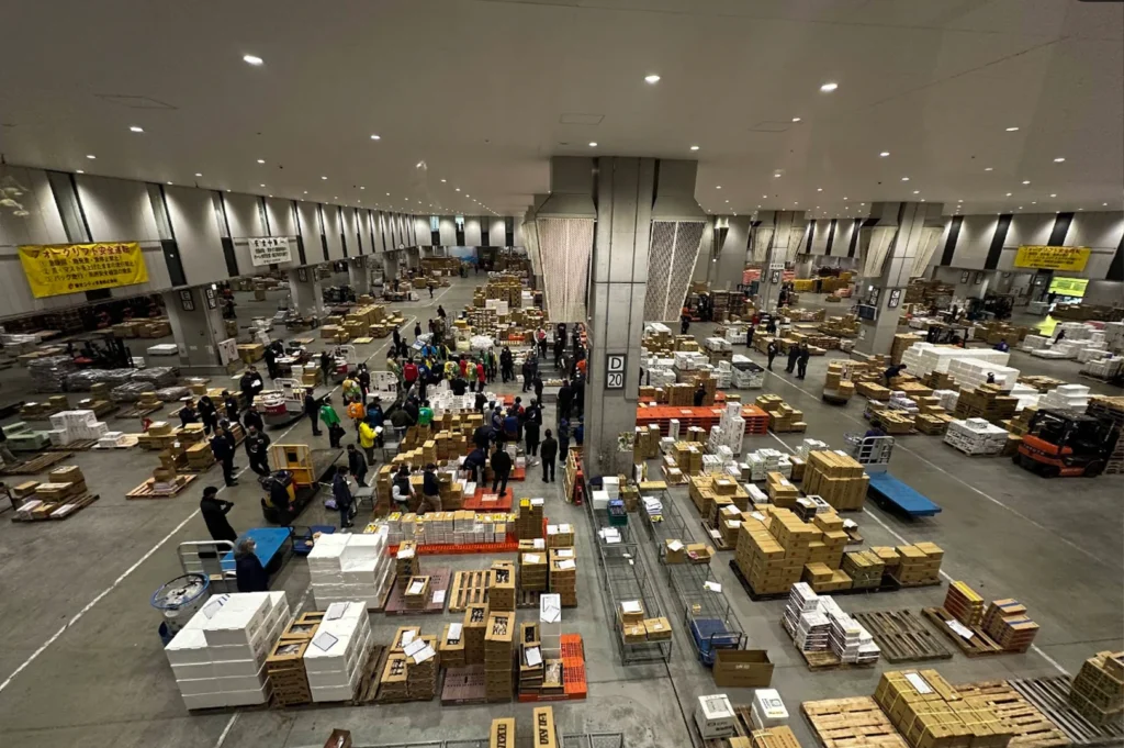 A wide-angle, birds-eye view reveals the massive scale of the Toyosu Market's wholesale hall. The vast, high-ceilinged room is filled with hundreds of stacked cardboard boxes and white styrofoam containers organized on pallets. Workers in various colored vests and caps move through the aisles, some gathered in small groups for auctions and others operating small motorized carts or forklifts. The industrial space is brightly lit by recessed ceiling lights, with large concrete pillars supporting the structure, illustrating the logistical complexity of one of the world's largest fish markets.