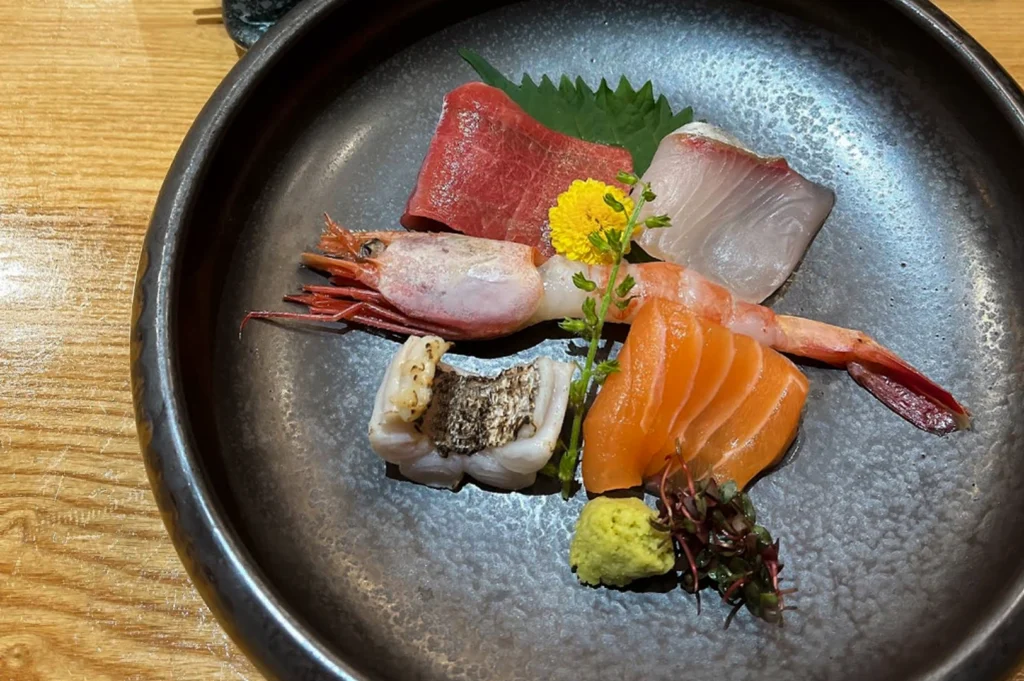 A top-down view of a dark ceramic bowl containing an assortment of fresh sashimi. The selection includes slices of salmon, tuna, white fish, a whole raw sweet shrimp (amaebi), and a piece of seared fish. It is garnished with a yellow chrysanthemum, a shiso leaf, wasabi, and purple sprouts.