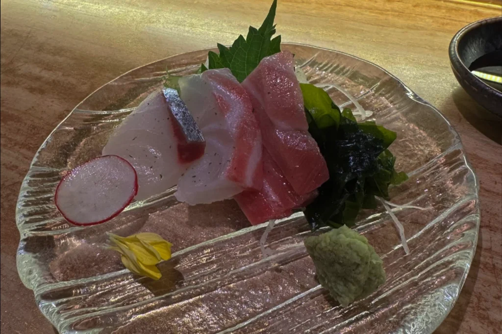 A top-down close-up shot showcases an elegant assortment of fresh sashimi beautifully arranged on a clear, textured glass plate. The plate holds thick, glistening slices of raw fish, including white-fleshed whitefish and rich pink fatty tuna with distinct marbling. The fish is garnished with a vibrant green shiso leaf, shredded white daikon radish, dark green seaweed, a single thin slice of red radish, and a small yellow flower petal. A neat mound of pale green wasabi rests at the bottom right corner of the plate.