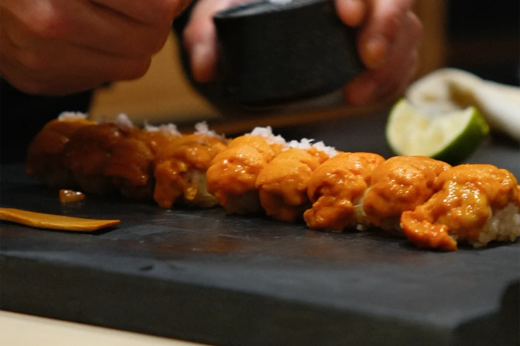 A chef's hands preparing a row of fresh Uni (sea urchin) nigiri sushi on a slate board, garnishing with sea salt.