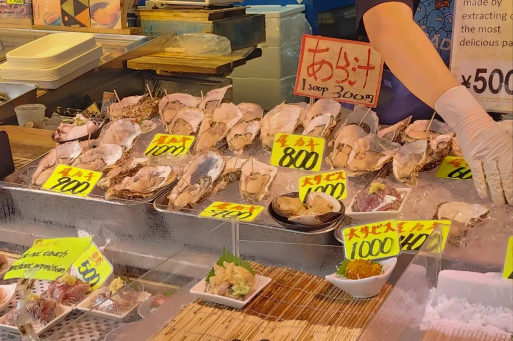 A close-up shot of a stainless steel tray filled with crushed ice, holding dozens of large, freshly shucked oysters in their craggy shells. Each oyster has a toothpick for easy eating. Bright yellow price tags are scattered across the ice, with prices like 800, 900, and 1,000 yen. In the foreground, small white rectangular dishes hold portions of sliced ginger and pale sashimi. To the right, a vendor wearing white latex gloves is partially visible, reaching toward the display.
