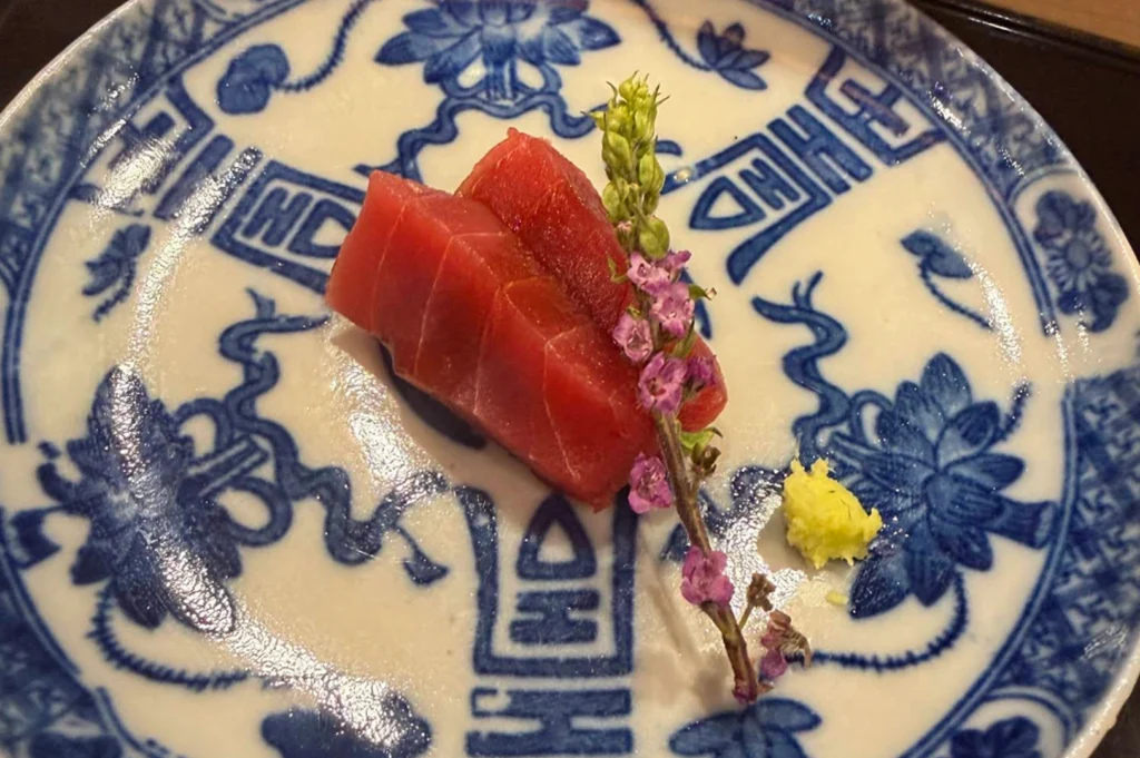 An artistic presentation of sashimi served on a decorative blue and white patterned ceramic plate. Two thick, rectangular slices of deep red tuna (maguro) are stacked slightly off-center. They are garnished with a delicate, upright sprig of purple shiso flowers and a small mound of bright yellow-green grated wasabi. The plate features traditional floral and geometric motifs in cobalt blue against a white background, giving the dish an elegant, classic feel.