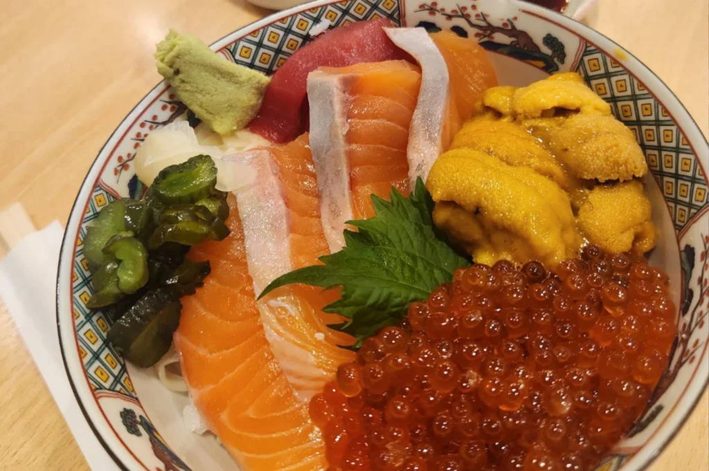 This close-up shot features a beautifully prepared "Kaisendon" or seafood bowl, served in a traditional patterned ceramic dish. The bowl is overflowing with premium raw ingredients: bright orange slices of salmon, deep red tuna (maguro), a generous mound of creamy yellow sea urchin (uni), and a large portion of glistening orange salmon roe (ikura). To the side, there are garnishes of bright green wasabi, pickled ginger, and dark green pickled cucumbers, all resting on a bed of sushi rice. The composition highlights the freshness and rich colors of the seafood, making for a highly appetizing culinary presentation.