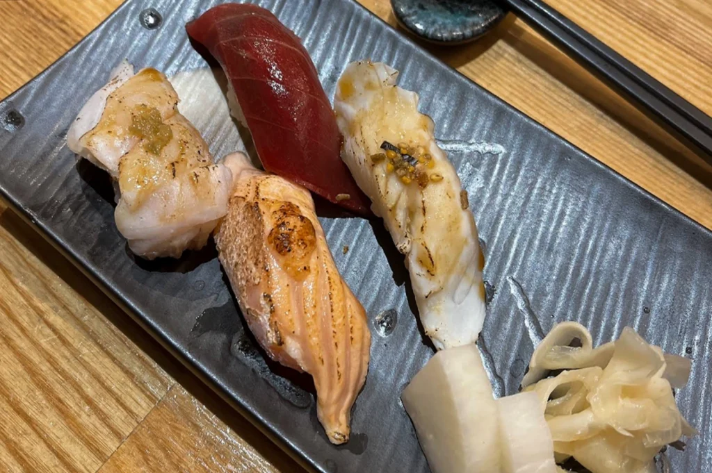 Four pieces of nigiri sushi served on a rectangular grey textured plate. The set features different toppings: seared white fish with yuzu kosho, a deep red tuna slice, seared salmon, and a piece of white fish with a savory topping. A side of pickled ginger and radish is included.