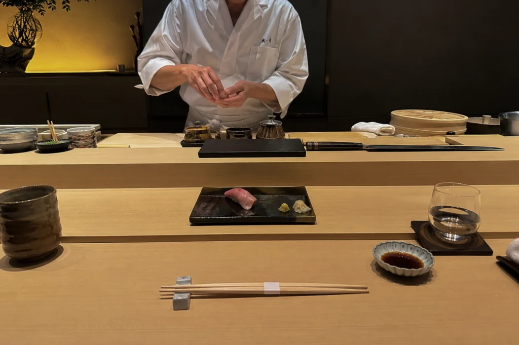 A front-facing medium shot captures a sushi chef standing behind a light wood counter, focused on shaping sushi with both hands. He wears a traditional white chef’s uniform with dark characters embroidered on the left chest. On the elevated wooden platform in front of him rests a single piece of tuna nigiri on a black ceramic plate, accompanied by a small pile of pickled ginger and wasabi. The lower counter features a pair of wooden chopsticks wrapped in a paper band on a ceramic rest, a small dish of soy sauce, a glass of water on a dark coaster, and a ceramic teacup.