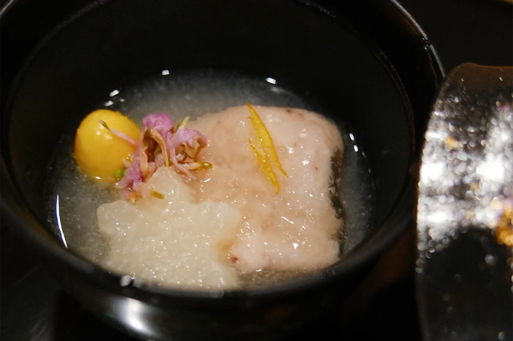 A delicate Japanese clear soup (Suimono) in a black lacquered bowl, featuring fish cake, a gingko nut, and yuzu zest.