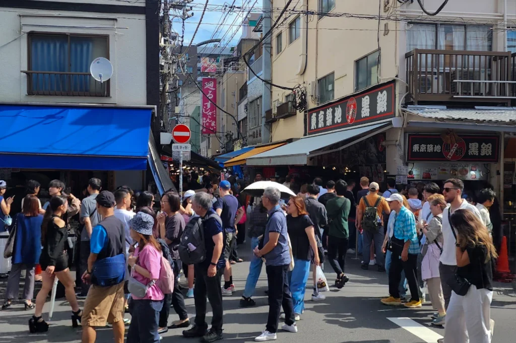A street-level view of a bustling, narrow intersection at the Tsukiji Outer Market. The street is packed with a diverse crowd of tourists and locals under a clear blue sky with many overhead power lines. To the right is a prominent shop with a black and red sign for "Tsukiji Kurogin," a famous tuna specialist, with a long line of people waiting underneath a white awning. The buildings are a mix of traditional and modern Japanese urban styles with blue and yellow awnings.