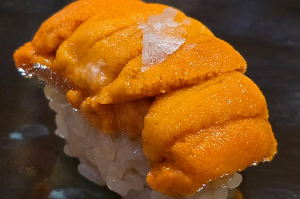 An extreme close-up of a piece of sea urchin (uni) nigiri, emphasizing its rich texture and vibrant color. The bright orange, creamy lobes of the uni are piled high atop a small bed of seasoned white sushi rice. A few large, translucent flakes of sea salt are sprinkled on the very top, catching the light. The background is a dark, matte surface that makes the warm tones of the seafood pop.