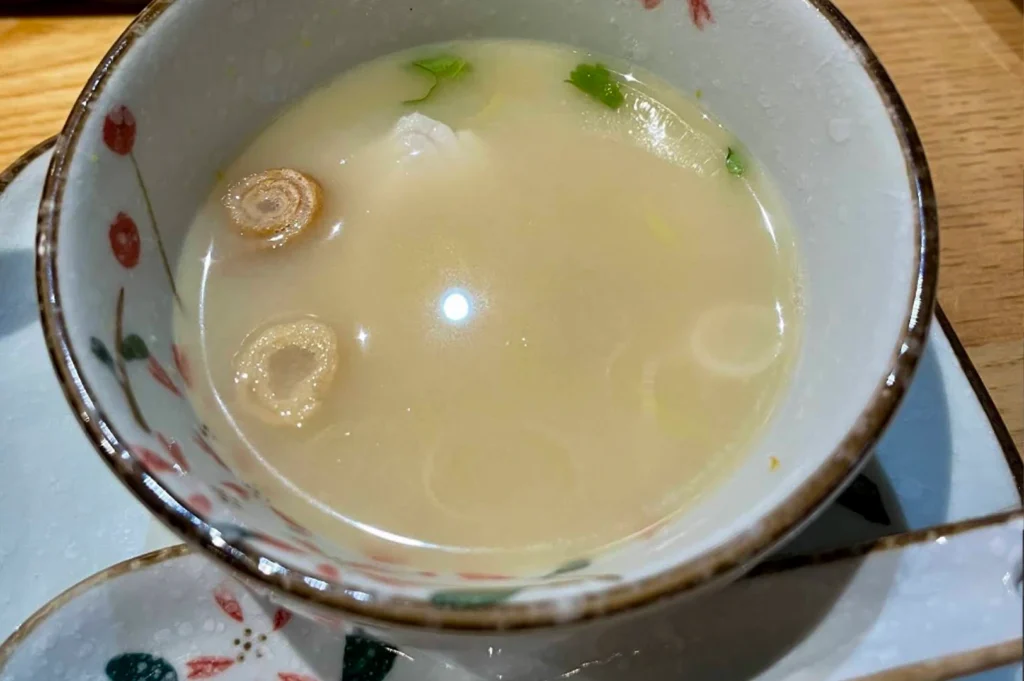 A close-up shot of a delicate, light-colored dashi soup served in a white ceramic bowl with pink floral patterns. The broth contains small pieces of green garnish and thin circular slices of Japanese leek.