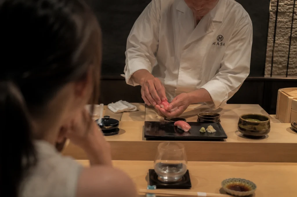 An eye-level medium shot shows a sushi chef behind a minimalist wooden counter, meticulously assembling a piece of nigiri sushi. He is wearing a crisp white chef's coat featuring a minimalist "M MASA" logo on the left chest. A female guest is captured from behind in the left foreground, watching his movements. On the black serving plate on the counter, one piece of pink tuna nigiri is already served alongside small portions of wasabi and ginger, while a glass of water and a small dish of soy sauce sit on the table below.