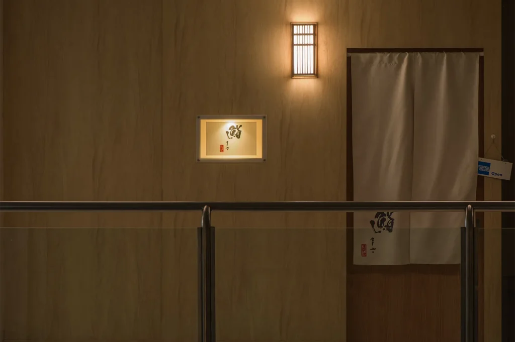The minimalist exterior entrance of a Japanese sushi restaurant with a white noren curtain and a glowing calligraphy sign.