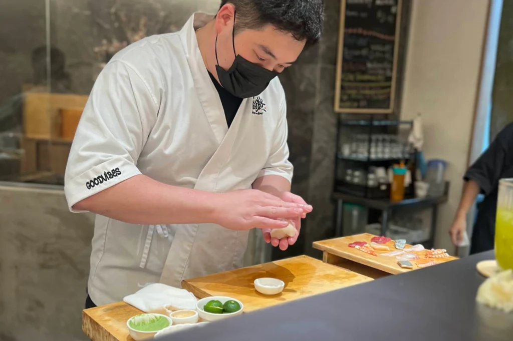 An eye-level action shot captures a Japanese sushi chef wearing a white jacket with "GOODVIBES" embroidered on the sleeve and a black face mask as he gently shapes a piece of nigiri with both hands. His wooden prep station features a platter of sliced fresh raw fish, small seasoning dishes containing wasabi and sudachi limes, and a wooden cutting board set against a modern restaurant kitchen backdrop.
