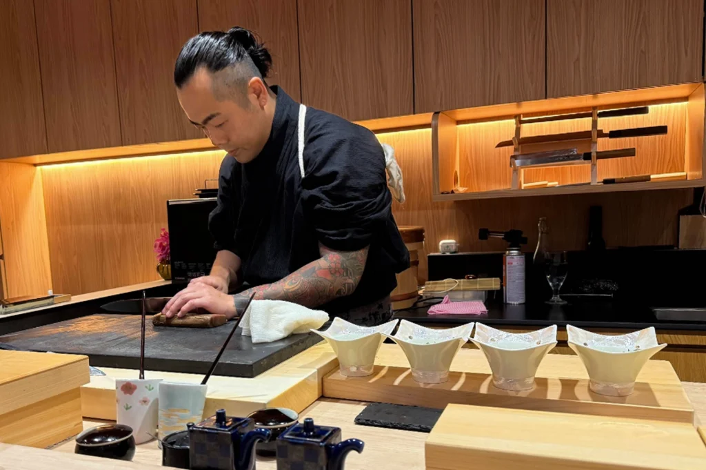 A wide shot of a sushi chef at work behind a clean, modern wooden counter. The chef, with his hair tied back and visible sleeve tattoos, is meticulously slicing a piece of fish on a dark stone workspace. In the foreground, a row of four elegant, white star-shaped ceramic bowls is lined up on a wooden riser, ready for the next course. The background features warm, recessed lighting illuminating wooden cabinetry and a display of professional sushi knives on a rack.