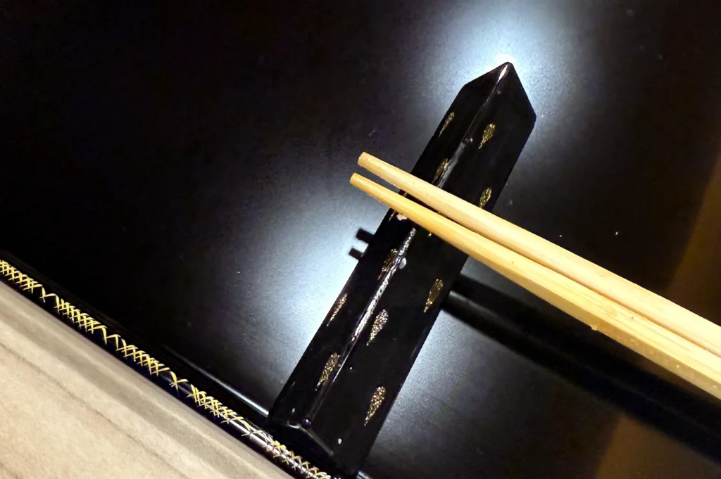Top-down view of wooden chopsticks resting on a black and gold decorated lacquer tray in a fine dining setting.