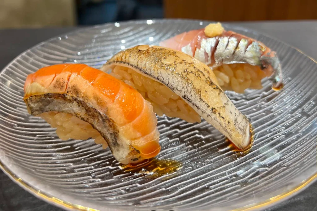 A close-up photograph displays three pieces of nigiri sushi arranged on a clear, textured glass serving dish with a subtle gold rim. The platter showcases a piece of salmon with lightly seared skin, a piece of torched white fish, and a piece of mackerel topped with grated ginger, all resting on akazu-seasoned rice drizzled with soy glaze.