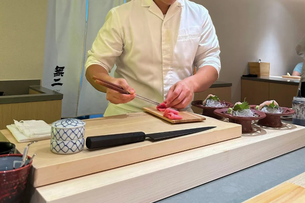 A sushi chef in a clean white uniform is captured in a mid-action shot behind a minimalist sushi bar, using long metal plating chopsticks to delicately fold and arrange slices of bright pink tuna. In front of him sits a thick, light-colored wooden cutting board holding a sleek black-handled sushi knife and a small blue-and-white patterned ceramic container. To the right, several finished sashimi platters are ready for service, each presented on a unique reddish-brown elevated stand with intricate latticework details.