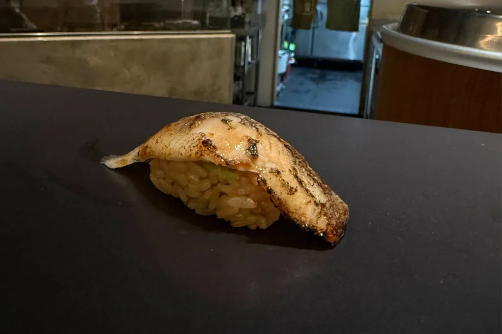 A close-up perspective focuses on a single piece of seared fish nigiri served directly onto a sleek dark counter surface. The fish skin is charred to a golden crispness, draped over a bed of brownish seasoned sushi rice under soft interior dining lights.