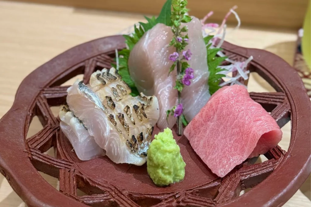 A beautifully arranged sashimi course is presented on a traditional reddish-brown ceramic stand with a decorative, star-shaped cutout pattern. The platter includes three distinct types of seafood: slices of translucent white fish, pieces of lightly seared fish with a charred skin texture, and a succulent fold of fatty pink tuna. The arrangement is garnished with a vibrant green shiso leaf, a vertical sprig of purple shiso flowers, delicate white radish curls, and a fresh mound of bright green wasabi in the center.