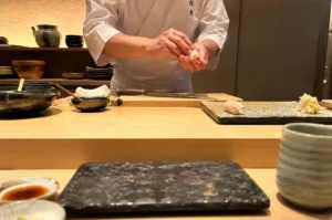 A first-person perspective from a diner's seat at an elegant, minimalist omakase sushi bar, focusing on a sushi chef in a traditional white uniform as they carefully shape a piece of nigiri sushi with their hands. In front of the chef on a light-colored wooden counter rests a large wooden cutting board with a long sushi knife, a small black bowl with a wooden brush, and a textured ceramic serving plate holding a single piece of white-fish nigiri and a small pile of pickled ginger. In the immediate foreground from the customer's point of view, a dark, mottled rectangular stone serving slab sits centered, flanked by a small dish of soy sauce on the left and a ceramic teacup on the right, all illuminated by warm, ambient lighting.