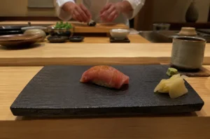A close-up, eye-level shot captures a single piece of premium tuna nigiri sushi resting in the center of a textured black rectangular slate plate at an omakase dining counter. The sushi features a glossy, finely seared slice of pinkish-red tuna draped over a small bed of seasoned rice, accompanied by a small pile of pickled ginger and a touch of green wasabi on the right side of the slate. In the softly blurred background, the skilled hands of a sushi chef in a white uniform are visible working at a light wooden prep station, surrounded by small ceramic bowls and fresh ingredients. The warm, low-key lighting and a rustic ceramic tea cup to the right enhance the intimate, authentic atmosphere of a traditional Japanese izakaya or high-end sushi bar.