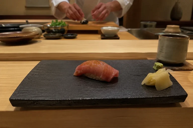 A close-up, eye-level shot captures a single piece of premium tuna nigiri sushi resting in the center of a textured black rectangular slate plate at an omakase dining counter. The sushi features a glossy, finely seared slice of pinkish-red tuna draped over a small bed of seasoned rice, accompanied by a small pile of pickled ginger and a touch of green wasabi on the right side of the slate. In the softly blurred background, the skilled hands of a sushi chef in a white uniform are visible working at a light wooden prep station, surrounded by small ceramic bowls and fresh ingredients. The warm, low-key lighting and a rustic ceramic tea cup to the right enhance the intimate, authentic atmosphere of a traditional Japanese izakaya or high-end sushi bar.