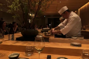 A professional male chef wearing a traditional white uniform and tall white chef's hat is meticulously preparing food behind a long, multi-tiered light wood counter inside a dimly lit, high-end Japanese restaurant. A second chef, also in white attire, stands slightly behind him to the left, focused on food preparation. The foreground features empty small bowls and glasses, including a glass filled with white wine, arranged on the smooth wooden surface. To the left, a large, dark textured stone pot holds a leafy green ornamental tree that extends toward the warm, wood-paneled ceiling, next to a large sake bottle with a white and black label. In the background, seated patrons along the counter chat and dine under a soft, intimate ambiance.