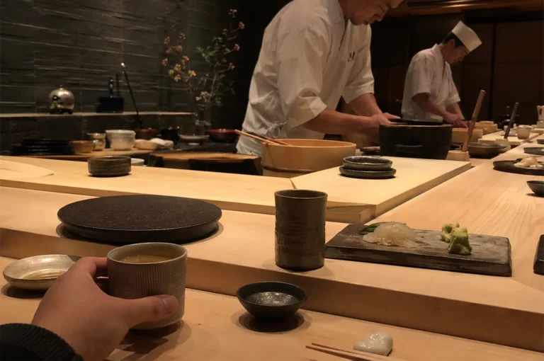 A first-person perspective captures the intimate atmosphere of a high-end sushi restaurant counter. In the foreground, a left hand holds a small ceramic cup filled with green tea, resting on a smooth, light-colored wooden bar alongside small dishes, a ceramic tumbler, a piece of prepared sushi served on a slate tile next to wasabi, and a set of chopsticks on a white ceramic rest. Across the wooden counter, two sushi chefs wearing traditional white coats and white caps are meticulously working behind the food preparation area, surrounded by wooden mixing bowls and ceramic plates. The background features a dark, textured stone tile wall subtly illuminated by warm overhead lighting, with a minimalist vase of delicate flowering branches adding an elegant touch to the moody, upscale ambiance.