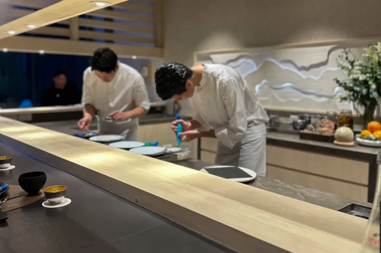A wide shot captures a clean, sophisticated omakase restaurant interior with two chefs in white uniforms working behind a long, polished light-wood counter. The foreground shows small, elegant tea bowls on a dark surface, while in the background, a chef uses a blue piping tool to carefully prepare a dish, creating an atmosphere of calm focus and dedicated craftsmanship.