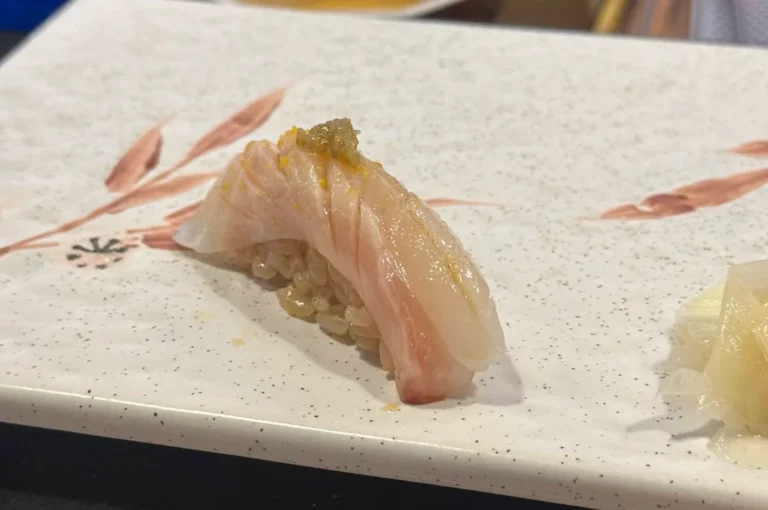 A close-up captures a single piece of nigiri sushi elegantly positioned on a speckled white ceramic plate decorated with subtle, painted brown leaves. The sushi consists of a slice of glossy, translucent white fish with delicate pink undertones, finely scored across the top and resting on a small bed of seasoned, brownish sushi rice. A tiny dab of green relish or seasoning sits perfectly on the crown of the fish, while a small pile of pickled ginger is visible in the lower right corner of the plate.