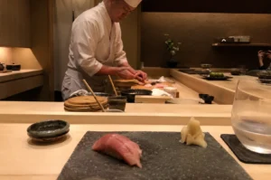 A close-up, first-person perspective at a minimalist Japanese sushi counter highlights a freshly prepared piece of tuna (maguro) nigiri resting on a dark slate serving block, accompanied by a small mound of pickled ginger (gari) and a glass of water to the right. In the soft-focused background, an expert sushi chef wearing a traditional white uniform and tall chef's hat (toque) leans intently over a light wooden counter, meticulously shaping sushi with his hands. The warm, intimate ambiance of the restaurant is accentuated by earthy, textured walls, subtle lighting, and various traditional ceramic dishes and wooden utensils neatly arranged across the counter space.
