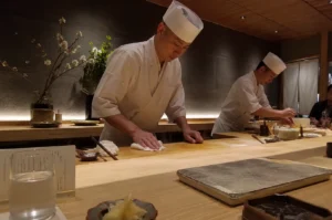 A high-angle, medium shot captures two sushi chefs in traditional white uniforms and tall chef hats working behind a light-colored wooden omakase counter in a dimly lit, minimalist restaurant. In the foreground, the primary chef stands centered, leaning forward slightly as he meticulously wipes down the pristine wooden surface with a white cloth, his expression one of quiet concentration. To his right, a second chef is partially visible further down the counter, focused on preparing ingredients. The setting exudes a refined, serene atmosphere with a textured, dark gray accent wall behind them, accented by subtle under-counter warm lighting and a minimalist floral arrangement of white blossoms in a dark vase to the left. On the customer's side of the bar, elements of the dining experience are visible in soft focus, including a glass of water, a small dish of pickled ginger, and a stone serving platter, emphasizing the intimate and attentive nature of the meal.
