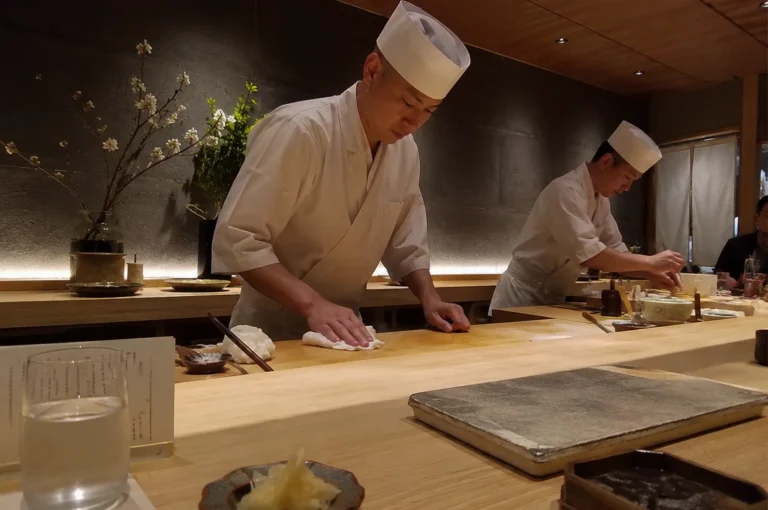 A high-angle, medium shot captures two sushi chefs in traditional white uniforms and tall chef hats working behind a light-colored wooden omakase counter in a dimly lit, minimalist restaurant. In the foreground, the primary chef stands centered, leaning forward slightly as he meticulously wipes down the pristine wooden surface with a white cloth, his expression one of quiet concentration. To his right, a second chef is partially visible further down the counter, focused on preparing ingredients. The setting exudes a refined, serene atmosphere with a textured, dark gray accent wall behind them, accented by subtle under-counter warm lighting and a minimalist floral arrangement of white blossoms in a dark vase to the left. On the customer's side of the bar, elements of the dining experience are visible in soft focus, including a glass of water, a small dish of pickled ginger, and a stone serving platter, emphasizing the intimate and attentive nature of the meal.