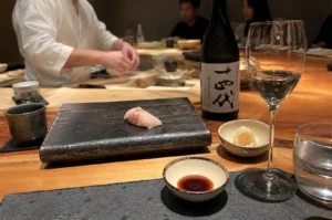 A close-up shot from the customer's perspective at a dimly lit, polished wooden sushi counter features a single piece of pale pink nigiri sushi topped with a dab of wasabi, presented on a dark, textured rectangular stone plate. To the right of the plate rests a small ceramic dish containing pickled ginger, a stemmed wine glass filled with clear sake, a clear water glass, and a small dipping dish filled with dark soy sauce. In the background, a bottle of Japanese sake with large black kanji calligraphy on a white label stands next to the wine glass, while the blurred figure of a sushi chef in a white uniform works at the counter, with other diners faintly visible in the soft, warm ambient lighting.