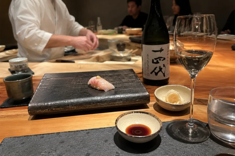 A close-up shot from the customer's perspective at a dimly lit, polished wooden sushi counter features a single piece of pale pink nigiri sushi topped with a dab of wasabi, presented on a dark, textured rectangular stone plate. To the right of the plate rests a small ceramic dish containing pickled ginger, a stemmed wine glass filled with clear sake, a clear water glass, and a small dipping dish filled with dark soy sauce. In the background, a bottle of Japanese sake with large black kanji calligraphy on a white label stands next to the wine glass, while the blurred figure of a sushi chef in a white uniform works at the counter, with other diners faintly visible in the soft, warm ambient lighting.