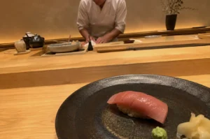 A single piece of tuna nigiri sushi is presented in the foreground on a dark, textured stoneware plate, accompanied by a small dollop of green wasabi and pickled ginger. The plate sits on a smooth, light-colored wooden counter that leads the eye toward the background, where a sushi chef wearing a white uniform is meticulously preparing food. The warm, minimalist setting features soft lighting bouncing off a golden-toned wall, with various traditional pottery bowls, a soy sauce dispenser, and a slender vase holding thin branches arranged neatly on the tiered wooden workspace.