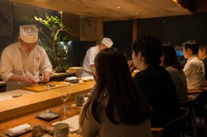 An intimate and warmly lit upscale sushi bar features a row of customers seated along a polished wooden counter, viewed from behind. On the left side of the counter, a professional sushi chef dressed in a traditional white uniform and chef's hat looks down intently as he meticulously prepares a dish on a wooden cutting board, while a second chef works diligently in the softly lit background next to a potted green bonsai tree. The patrons, including a couple in the foreground and several others down the line, watch the preparation in a relaxed atmosphere, with various small plates, a glass of white wine, tea cups, and folded wet towels neatly arranged along the counter before them under a warm wooden ceiling.