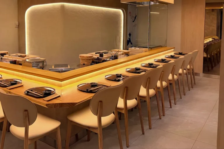 The warm, inviting interior of an omakase restaurant is showcased, featuring a long, polished light-wood counter with multiple place settings consisting of black plates, chopsticks, and small bowls, arranged neatly for diners. The space is characterized by light-wood chairs, clean floor tiles, and a minimalist design enhanced by soft, warm accent lighting along the bar and walls, creating an atmosphere of understated luxury and culinary focus.