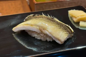 A piece of nigiri sushi featuring a slice of fish with a distinctive dark-to-light gradient on its skin is resting on a sleek, black rectangular plate. The fish is draped over a mound of rice, and a piece of light yellow pickled ginger is positioned on the right side of the plate, reflecting faintly on the dark, smooth surface.